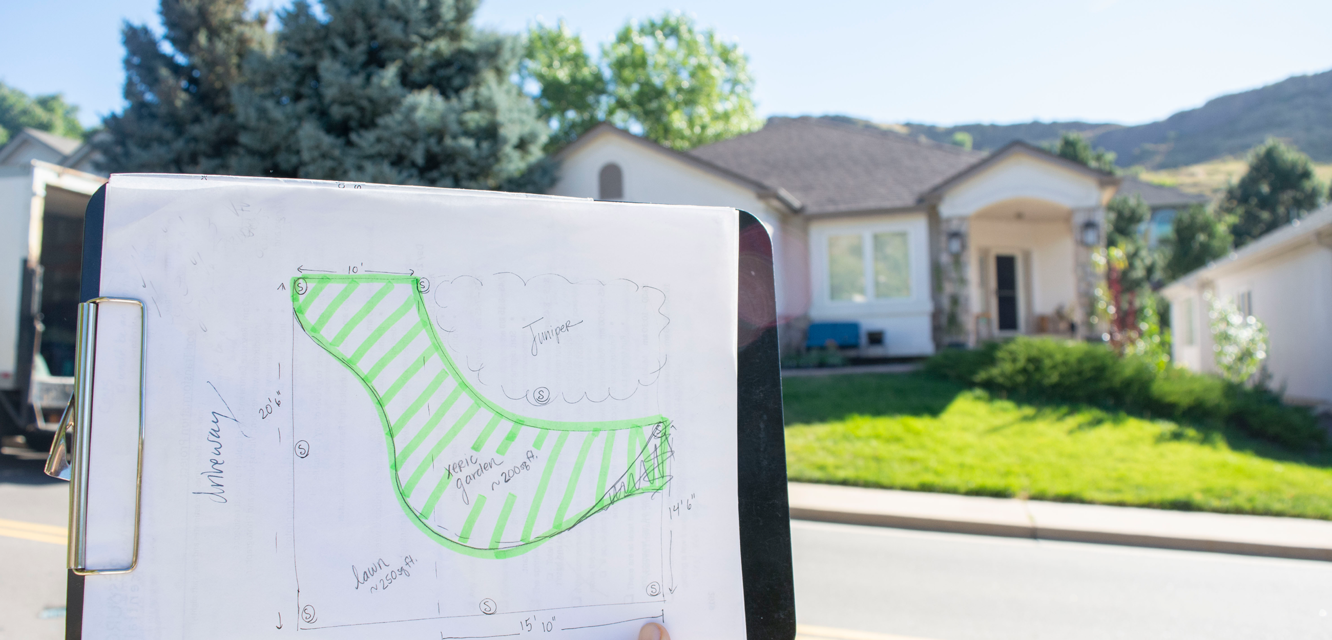 A clipboard holding a drawing of a landscape is held in front of the house with a green lawn